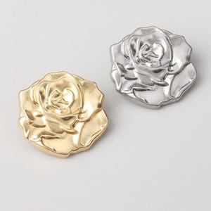 Puede incluir: Dos objetos decorativos metálicos con forma de rosa. Uno es de color dorado, el otro plateado. Las rosas están detalladas con pétalos y una superficie texturizada. Los objetos probablemente se utilizan para embellecer o decorar.