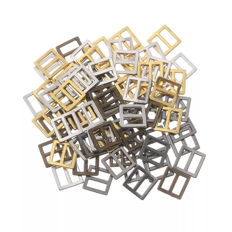 Op de afbeelding: Een stapel metalen schuifgespen in gouden, zilveren en bronzen afwerkingen. De gespen zijn vierkant met een rechthoekige opening in het midden.