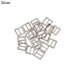 Op de afbeelding: Een stapel zilverkleurige metalen schuifgespen. De gespen zijn rechthoekig met een kleine opening in het midden.