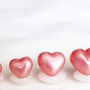 May include: Four pink heart-shaped buttons of varying sizes, arranged in a row from smallest to largest. The buttons are made of a shiny, pearlescent material and are mounted on white bases.