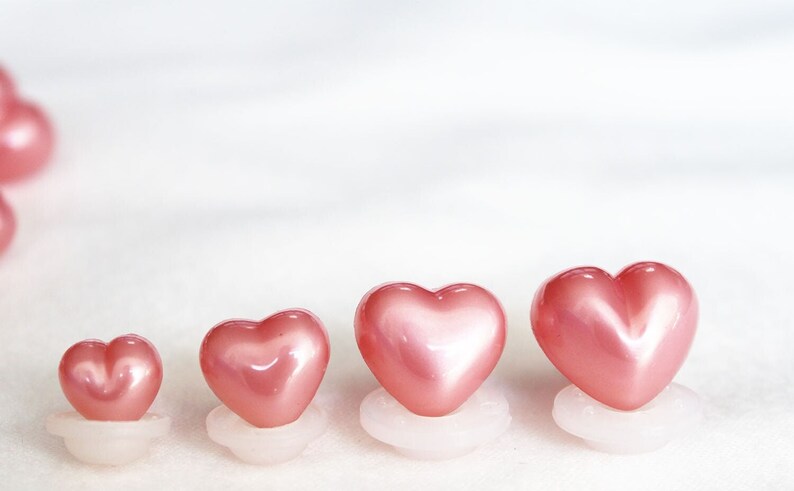 May include: Four pink heart-shaped buttons of varying sizes, arranged in a row from smallest to largest. The buttons are made of a shiny, pearlescent material and are mounted on white bases.