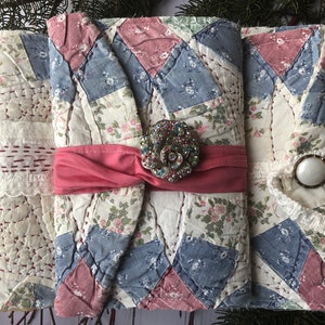 May include: Three handmade fabric journals with a patchwork design in shades of blue, pink, and white. Each journal has a different embellishment, including a jeweled flower, a white button, and a black button.