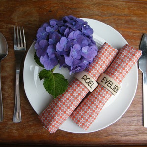 May include: Two orange and white patterned napkins with wooden name card holders that say "Roel" and "Evelien". The napkins are on a white plate with a purple hydrangea flower.