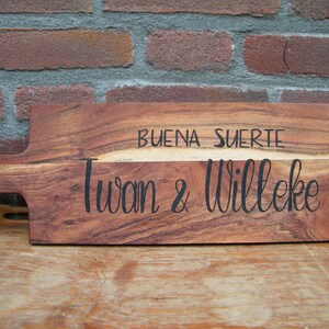 May include: A wooden cutting board with a handle, engraved with the text "Buena Suerte Iwan & Willeke".
