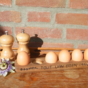 May include: Wooden egg holder with the inscription "Tout Van Bien" and a floral design. The holder has five slots for eggs and is sitting on a wooden surface. Two wooden salt and pepper shakers are in the background.