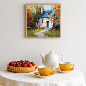 Op de afbeelding: Een ingelijst schilderij van een schilderachtig huisje met een blauw dak en een turkooizen deur hangt boven een tafel. Op de tafel staan een aardbeientaart en twee gele theekopjes met schotels, samen met een witte theepot.