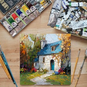 Op de afbeelding: Een olieverfschilderij van een huisje met een blauw dak en een blauwe deur, omringd door kleurrijk gebladerte. Verftubes, penselen en kunstbenodigdheden zijn rondom het schilderij op een houten oppervlak geplaatst.