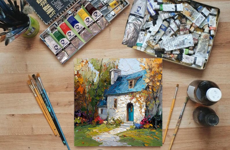 Op de afbeelding: Een olieverfschilderij van een huisje met een blauw dak en een blauwe deur, omringd door kleurrijk gebladerte. Verftubes, penselen en kunstbenodigdheden zijn rondom het schilderij op een houten oppervlak geplaatst.