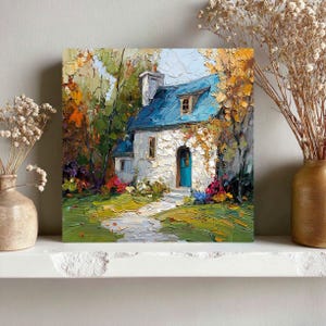 Op de afbeelding: Vierkant olieverfschilderij van een schilderachtig huisje met een blauw dak en deur, omringd door bomen met herfstkleuren. Een stenen pad leidt naar de ingang. Het kunstwerk wordt tentoongesteld op een witte plank met gedroogde bloemen in vazen.