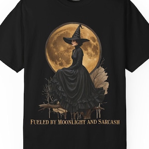 May include: Black t-shirt featuring a witch in a black dress and hat, seated in front of a full moon. The text "FUELED BY MOONLIGHT AND SARCASM" is printed below the image. The shirt is made of a soft material.