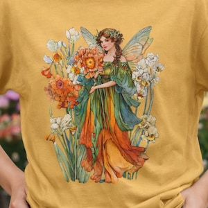 Puede incluir: Camiseta de color amarillo mostaza con una ilustración de una hada sosteniendo flores. El hada lleva un vestido verde y naranja con alas azules. Las flores son naranjas, blancas y amarillas. La camiseta es de material suave.
