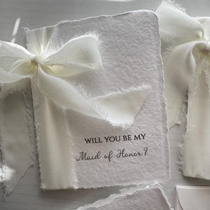 May include: Ivory-colored invitation cards with deckled edges, tied with a delicate white ribbon. The text "WILL YOU BE MY Maid of Honor?" is printed in elegant script. Ideal for wedding proposals.