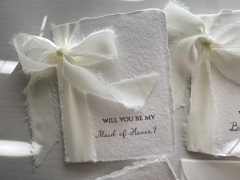 May include: Ivory-colored invitation cards with deckled edges, tied with a delicate white ribbon. The text "WILL YOU BE MY Maid of Honor?" is printed in elegant script. Ideal for wedding proposals.