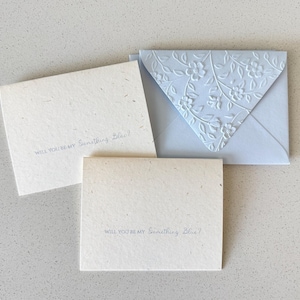 Könnte beinhalten: Drei Schreibwarenartikel: zwei cremefarbene Karten und ein hellblauer Umschlag. Auf den Karten steht in zarter blauer Schrift "WILL YOU BE MY Something Blue?". Der Umschlag hat ein geprägtes Blumendesign.