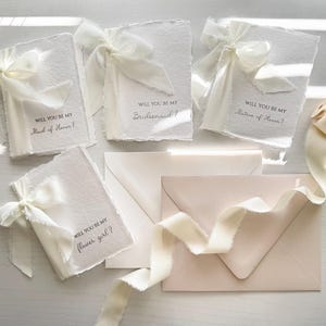 May include: Ivory-colored invitation cards with torn edges, tied with silk ribbons. The cards read "Will you be my Maid of Honor?", "Bridesmaid?", "Matron of Honor?", and "Flower girl?" Several matching envelopes and a spool of ribbon are also present.
