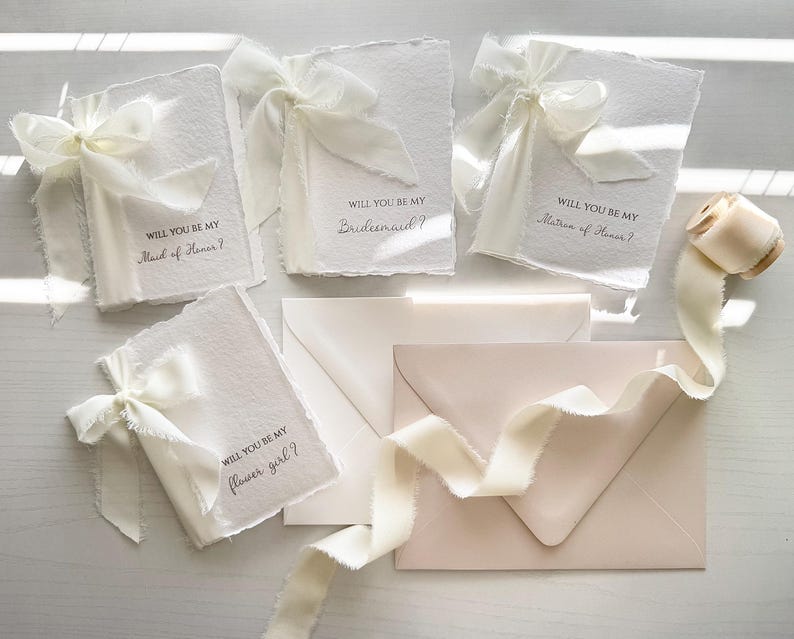 May include: Ivory-colored invitation cards with torn edges, tied with silk ribbons. The cards read "Will you be my Maid of Honor?", "Bridesmaid?", "Matron of Honor?", and "Flower girl?" Several matching envelopes and a spool of ribbon are also present.