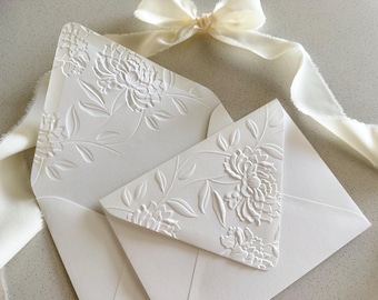 May include: Two cream-colored envelopes featuring an embossed floral pattern on the flaps. The envelopes are accompanied by a long, flowing cream-colored silk ribbon with frayed edges that is tied in a bow above the stationery set.