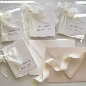 May include: White invitation cards with deckled edges, tied with ivory ribbons. Text on the cards reads "Will you be my Maid of Honor?", "Bridesmaid?", "Matron of Honor?", and "Flower Girl?". Envelopes and ribbon complete the wedding stationery set.