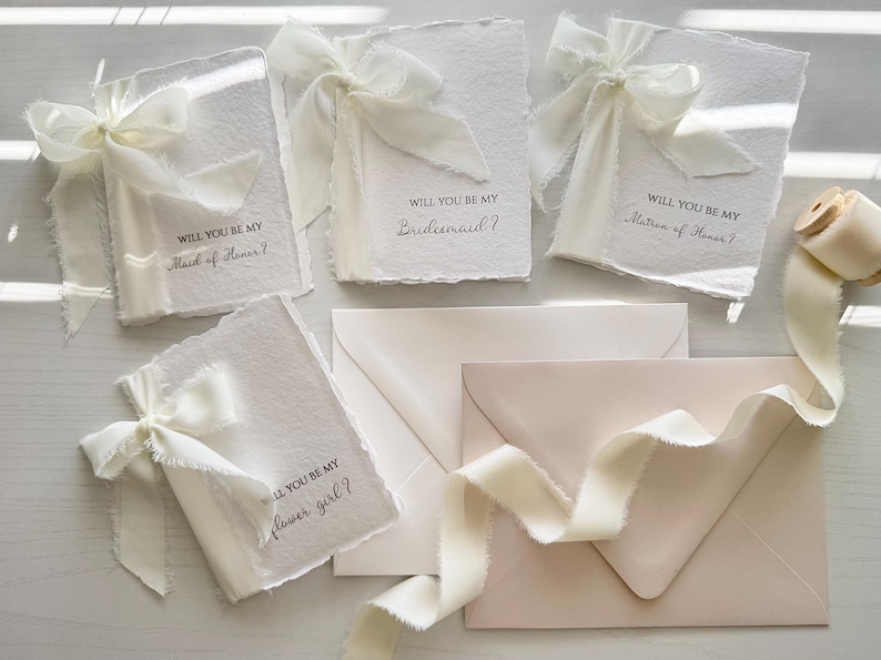 May include: White invitation cards with deckled edges, tied with ivory ribbons. Text on the cards reads "Will you be my Maid of Honor?", "Bridesmaid?", "Matron of Honor?", and "Flower Girl?". Envelopes and ribbon complete the wedding stationery set.