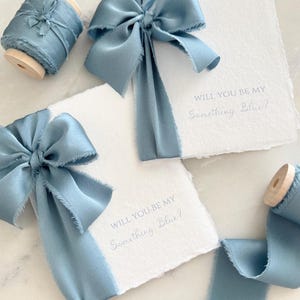 May include: Soft blue silk ribbon and bows accent white cards. The cards read "Will You Be My Something Blue?" A spool of matching ribbon is also present. The items are arranged on a light-colored surface, creating a delicate and romantic aesthetic.