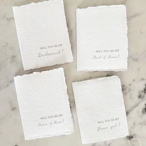 May include: Four white rectangular cards with deckled edges. Each card has the phrase "WILL YOU BE MY" followed by a different title: "Bridesmaid?", "Maid of Honor?", "Matron of Honor?", and "flower girl?" in elegant script.