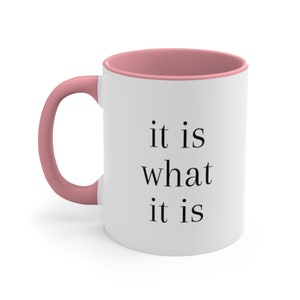 Può includere: Tazza in ceramica bianca con bordo e manico rosa. La tazza ha un testo nero che recita "it is what it is".