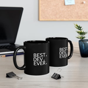 Puede incluir: Dos tazas de cerámica negra con texto blanco que dice "BEST. DEV. EVER." sobre una superficie de madera.