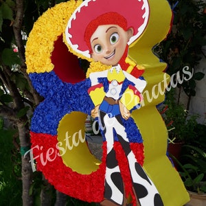 May include: A red, yellow, and blue pinata shaped like the number 8 with a cartoon character of a female cowboy wearing a red hat and a blue and white shirt. The pinata is decorated with tissue paper and has the text "Fiesta de Pi&ntilde;atas" on it.