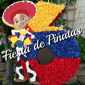 May include: A red, yellow, and blue pinata shaped like the number 5. The pinata is decorated with tissue paper and features a cartoon image of a female character wearing a cowboy hat and a red and yellow shirt. The text "Fiesta de Pi&ntilde;atas" is written in white on the pinata.