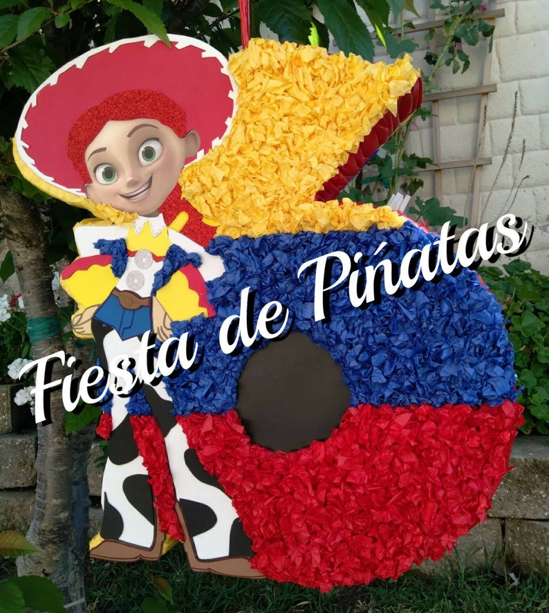 May include: A red, yellow, and blue pinata shaped like the number 5. The pinata is decorated with tissue paper and features a cartoon image of a female character wearing a cowboy hat and a red and yellow shirt. The text "Fiesta de Pi&ntilde;atas" is written in white on the pinata.