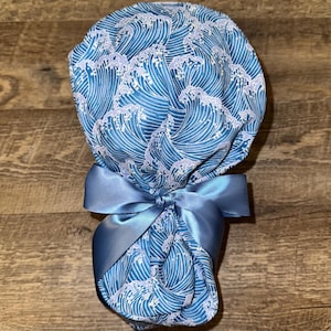 May include: A scrub cap featuring a repeating pattern of stylised blue and white ocean waves. The cap is gathered at the back and secured with a solid light blue satin ribbon tied into a bow, creating a rounded shape with a smooth fabric finish.