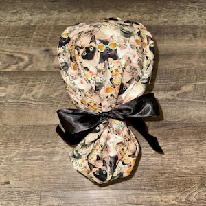 May include: A patterned scrub cap featuring a repeating design of cats wearing glasses and sunglasses. The cap is tied with a large black satin bow. The background is a light beige color.