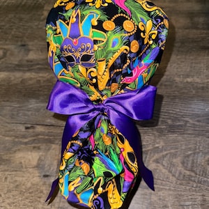 May include: A purple and gold fabric scrub hat with a Mardi Gras theme. The hat features a jester mask, peacock feathers, and gold coins. The hat is tied with a purple satin ribbon.
