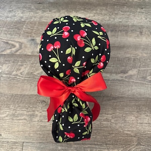 May include: A black fabric scrub cap with a red cherry and green leaf pattern, accented with white polka dots. A large red satin bow is tied at the base. The cap is designed for medical professionals.