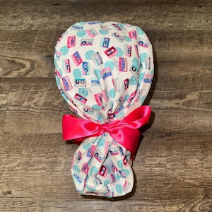May include: A white scrub cap featuring a pattern of pink and blue cassette tapes and teal polka dots. The cap is tied with a bright pink satin bow. The cap is designed for medical professionals.