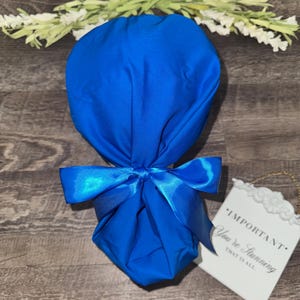 May include: A solid royal blue scrub cap featuring a gathered back design secured with a matching satin ribbon tie. The fabric is a vibrant, uniform blue color throughout, providing a clean and functional head covering for medical or clinical use.