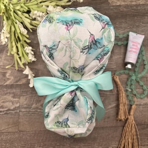May include: A scrub cap featuring a watercolor print of hummingbirds and green foliage on a white background. The cap is gathered at the back and secured with a bright mint green satin ribbon tied into a bow, creating a ponytail-style silhouette.