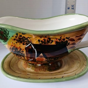 May include: A green and brown ceramic gravy boat with a painted scene of a house and trees. The boat has a matching saucer.