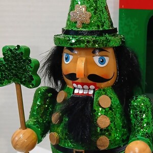 May include: An 8-inch nutcracker figure wearing a green glittery hat with a gold shamrock, a matching green glittery jacket with gold buttons, and black boots. The figure holds a green shamrock staff and a black pot filled with gold coins.