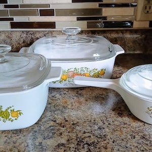 May include: Three white ceramic cookware pieces with clear glass lids. The square dish is labeled L'Echalote, the rectangular dish is labeled La Marjolaine, and the round saucepan is labeled Le Persil, all featuring colorful vegetable motifs.