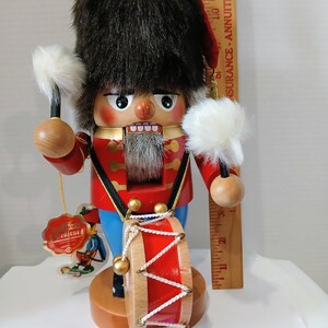 May include: A wooden nutcracker soldier wearing a tall brown faux fur hat and a red jacket with gold trim, holding two white drumsticks. A red drum is attached to the front. A wooden ruler shows the figure is approximately 12 inches tall.