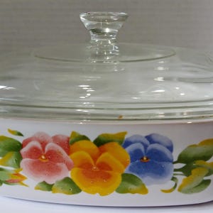 May include: A rectangular white ceramic casserole dish with a clear glass lid. The exterior features a painted floral design of pink, yellow, and blue pansies with green leaves. The dish has integrated white handles on both sides.
