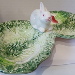May include: A ceramic serving dish shaped like a cabbage leaf, with a white bunny figurine perched on top. The dish is green with darker green veining details. The bunny has pink accents and red eyes.