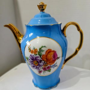 May include: A light blue porcelain teapot with a gold handle, spout, and accents. The teapot features a floral design with orange, purple, and pink flowers on a white oval. The lid is light blue with a gold finial.