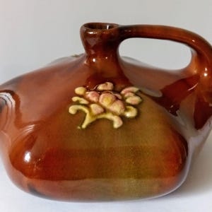 May include: A brown ceramic jug with a handle and a decorative floral design. The jug has a glossy finish and a unique shape, suitable for display or use. The floral design is in shades of yellow and pink.