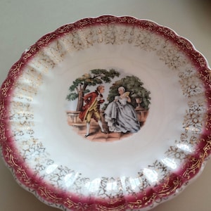 May include: A white ceramic bowl with a scalloped edge, featuring a burgundy and gold floral design. The center depicts a couple in period clothing near a tree. A decorative piece for serving or display.
