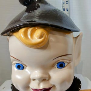 May include: A vintage ceramic cookie jar shaped like a smiling elf with blue eyes, a brown hat, and a red mouth. The elf has blonde hair and large ears. The jar is a collectible item.