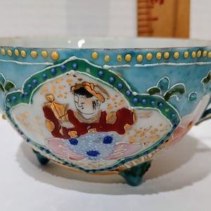 May include: A turquoise porcelain teacup with a handle and three small feet. The cup features a hand-painted design of a person in traditional clothing, with gold and red accents. The rim is decorated with gold and blue details.