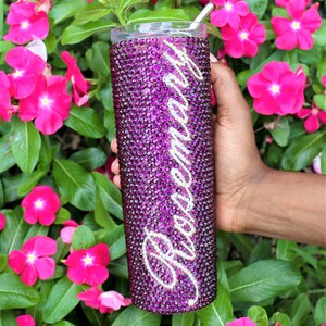 May include: A purple tumbler with a silver straw and the word "Remarkable" in silver rhinestones. The tumbler is covered in purple rhinestones.