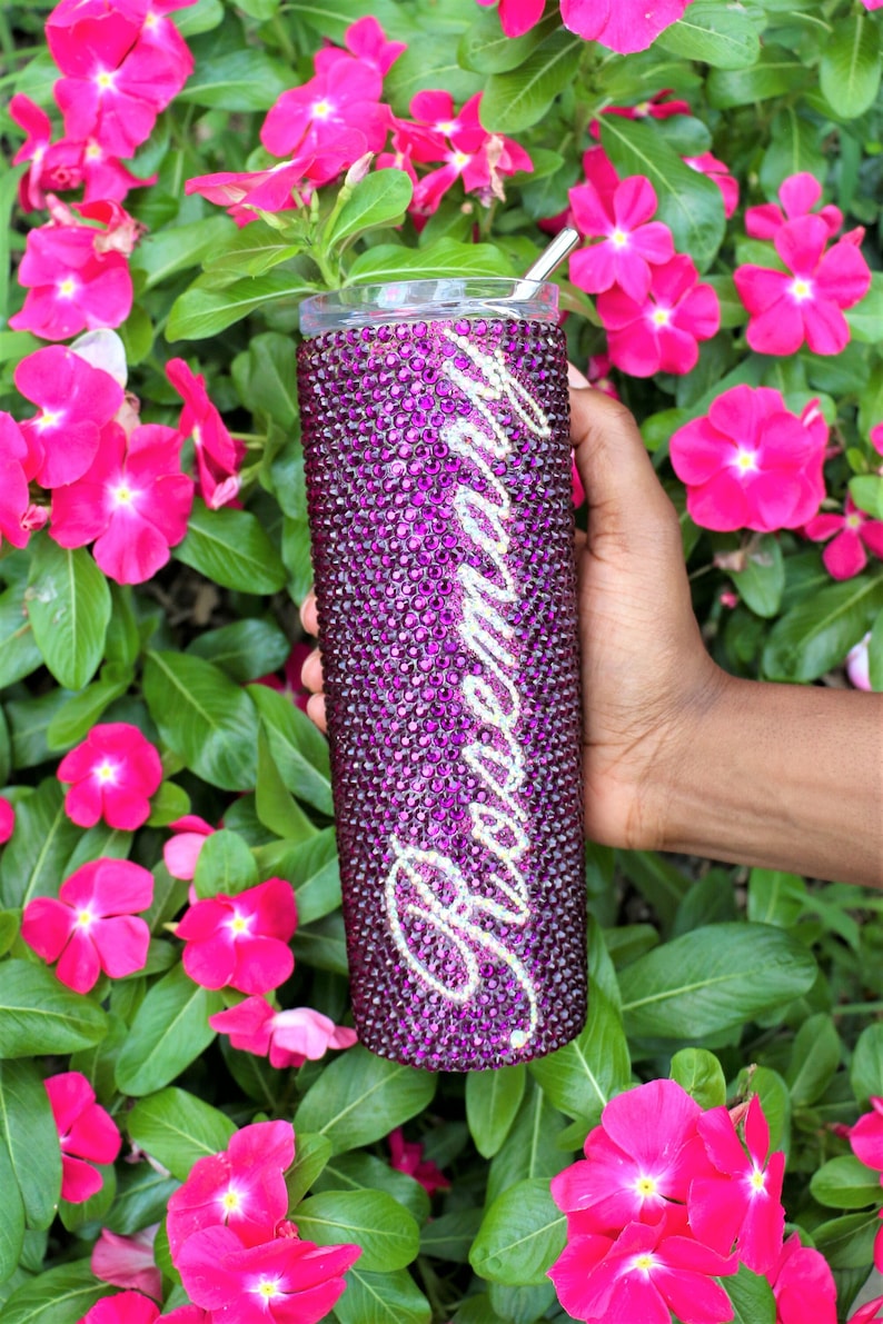 May include: A purple tumbler with a silver straw and the word "Remarkable" in silver rhinestones. The tumbler is covered in purple rhinestones.